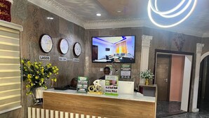 A hotel reception area with a wooden reception desk, a television displaying a room, and clocks labeled with city names. - Marchforte-Courtyard Hotel  (Ibadan)