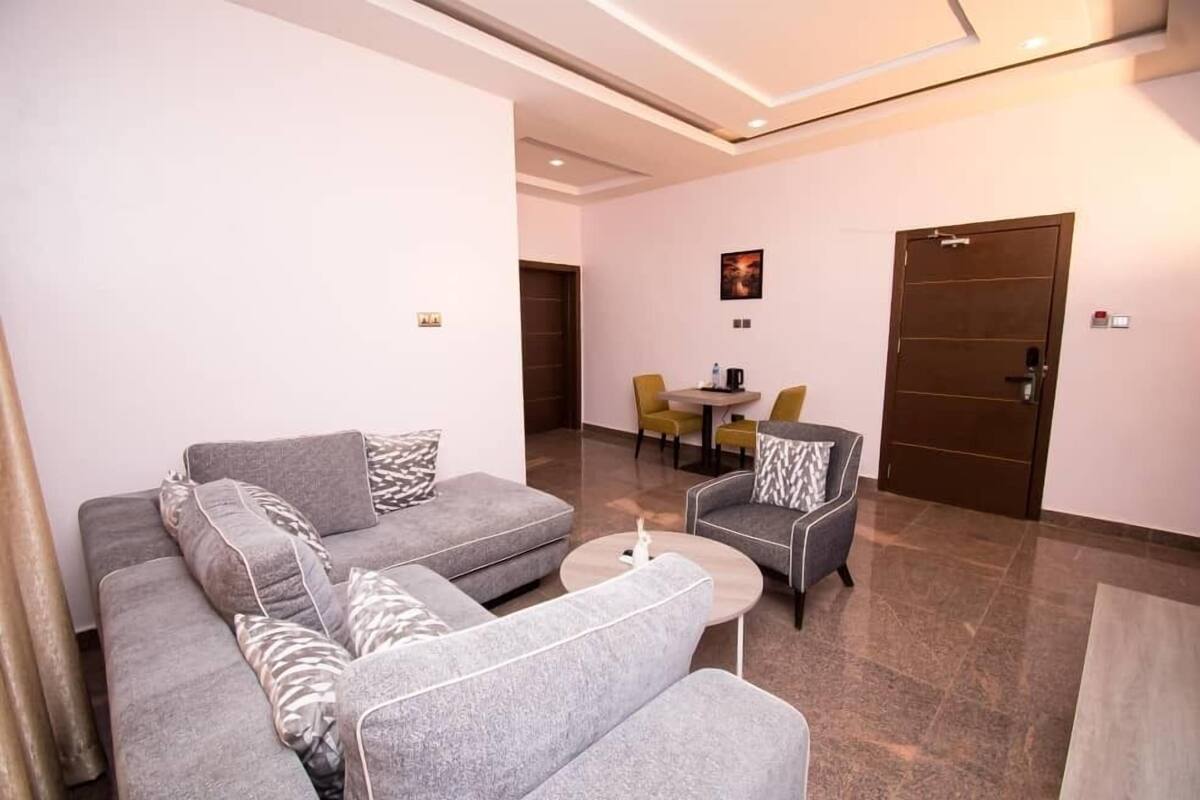 Een moderne woonkamer met grijze banken en fauteuils, een salontafel, en een eethoek met gele stoelen.