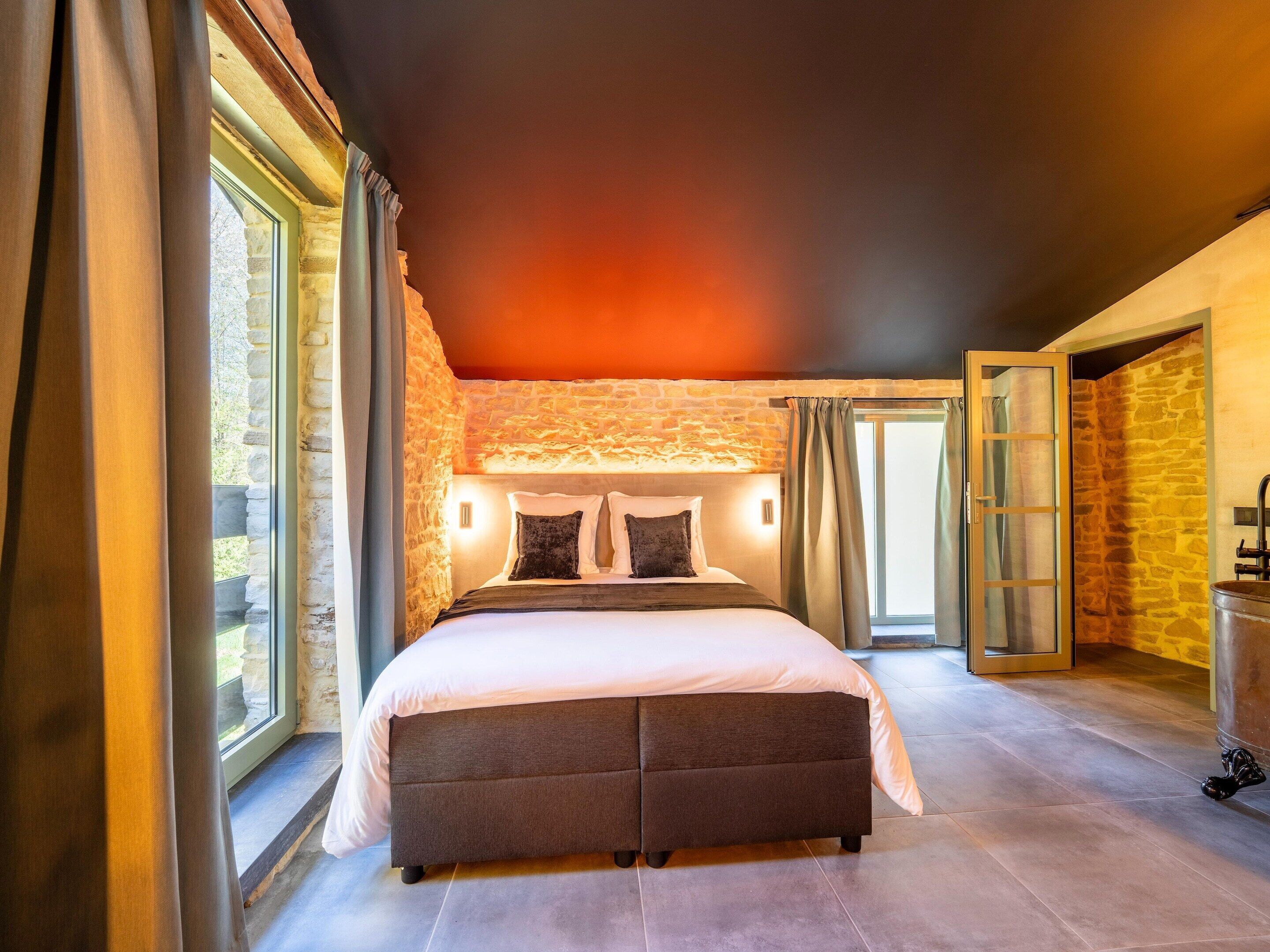 A bedroom with a stone wall, a large bed, a wooden ceiling, and a view through a window.