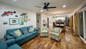 Ein Wohnzimmer mit einem blauen Sofa, zwei Sesseln, Holzboden, einem Deckenventilator und einer Küche im Hintergrund.