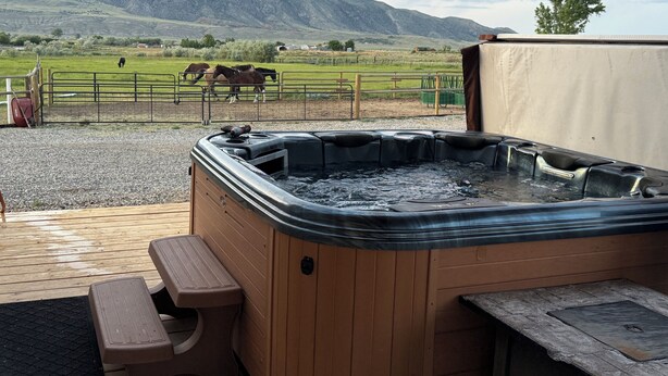 A hot tub with a wooden deck and a view of horses grazing in a fenced field with mountains in the background.