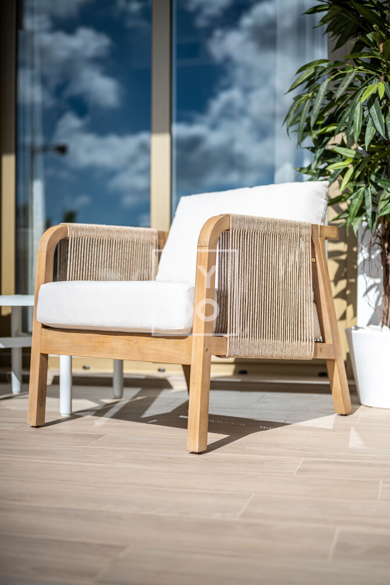 A wooden lounge chair with a woven backrest and white cushion, positioned on a tiled patio with a potted plant nearby.