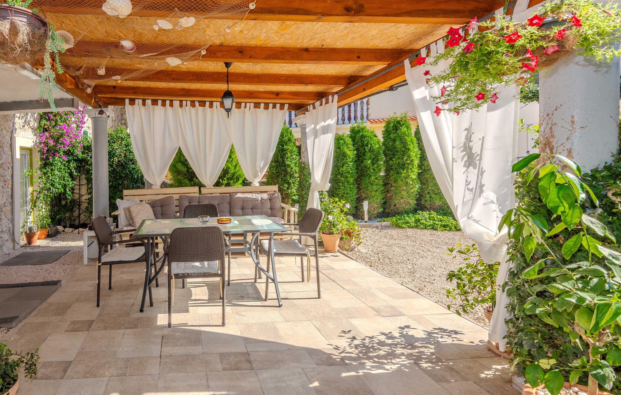En udendørs terrasse med et træbeklædt pergolaoverdækning, et spisebord med stole og et loungeområde med hynder.