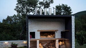 An outdoor stone fireplace with a wooden mantel, surrounded by a stone wall and a grassy area.