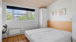 Un dormitorio con cama, una silla, un radiador y una ventana que ofrece vistas a árboles y un edificio.