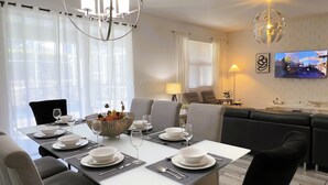 A modern dining area with a white table set for six, surrounded by grey upholstered chairs. A decorative basket with dried flowers is placed on the table.