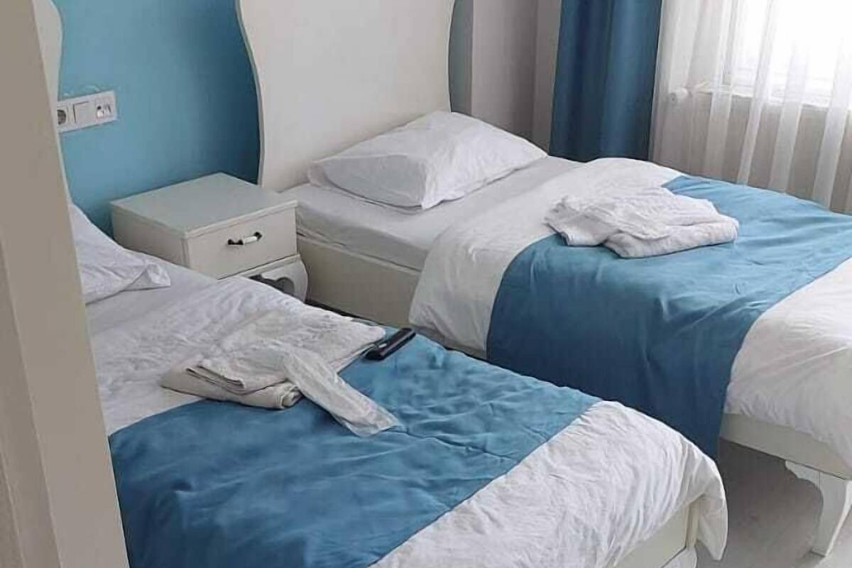 A room with two beds, each with white bedding and blue accents, a white nightstand, and a blue curtain.