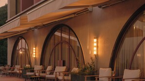 Un área de estar al aire libre con sillas y mesas, amplias ventanas en arco y un edificio de estilo arquitectónico moderno.