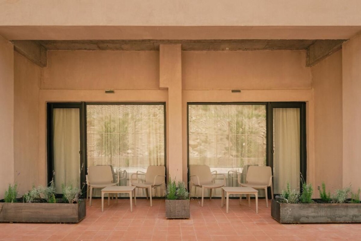 A covered outdoor seating area with two rows of chairs and tables, large windows with curtains, and planters with greenery.