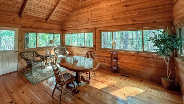 L’intérieur d’un chalet en bois comprend une table à manger ronde, des chaises, un ventilateur de plafond et de grandes fenêtres donnant sur la forêt.