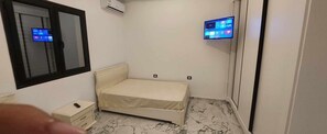A small, narrow room with a single bed, a bedside table, an air conditioning unit, and a wall-mounted television. - Villa hasdrubal swimming pool not overlooked (Djerba Midun)