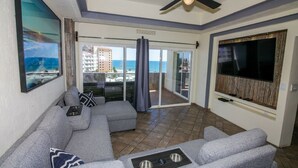 A living room with a gray sectional sofa, a ceiling fan, a TV, and a balcony with a view of the ocean and buildings. - Bonita 602 (Puerto Peñasco)