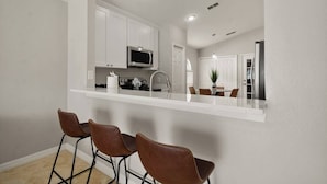 A modern kitchen with white cabinets, a microwave, and a sink. There are three brown chairs and a dining table.