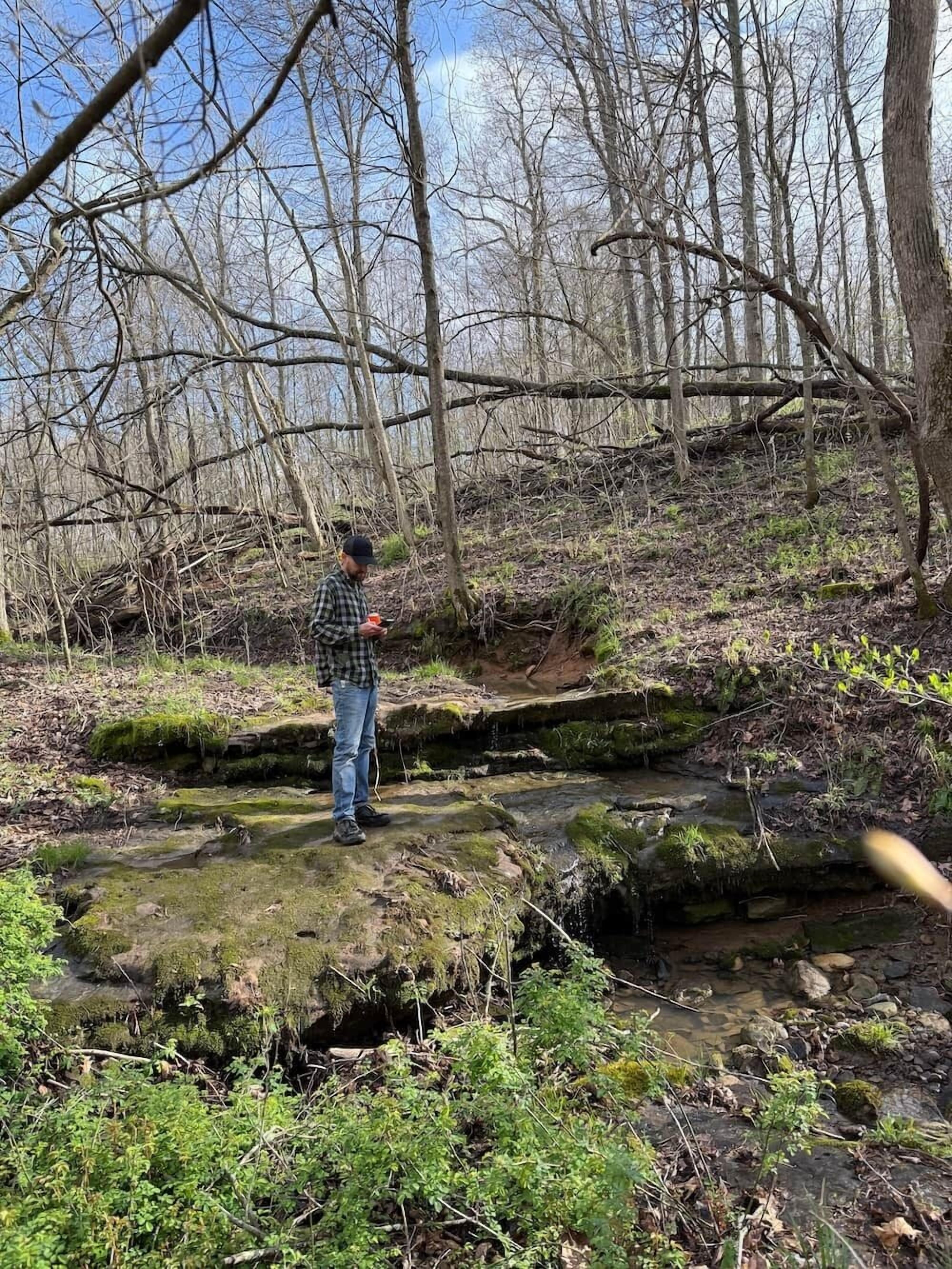 A person standing on a moss-covered rock in a forested area with a stream.