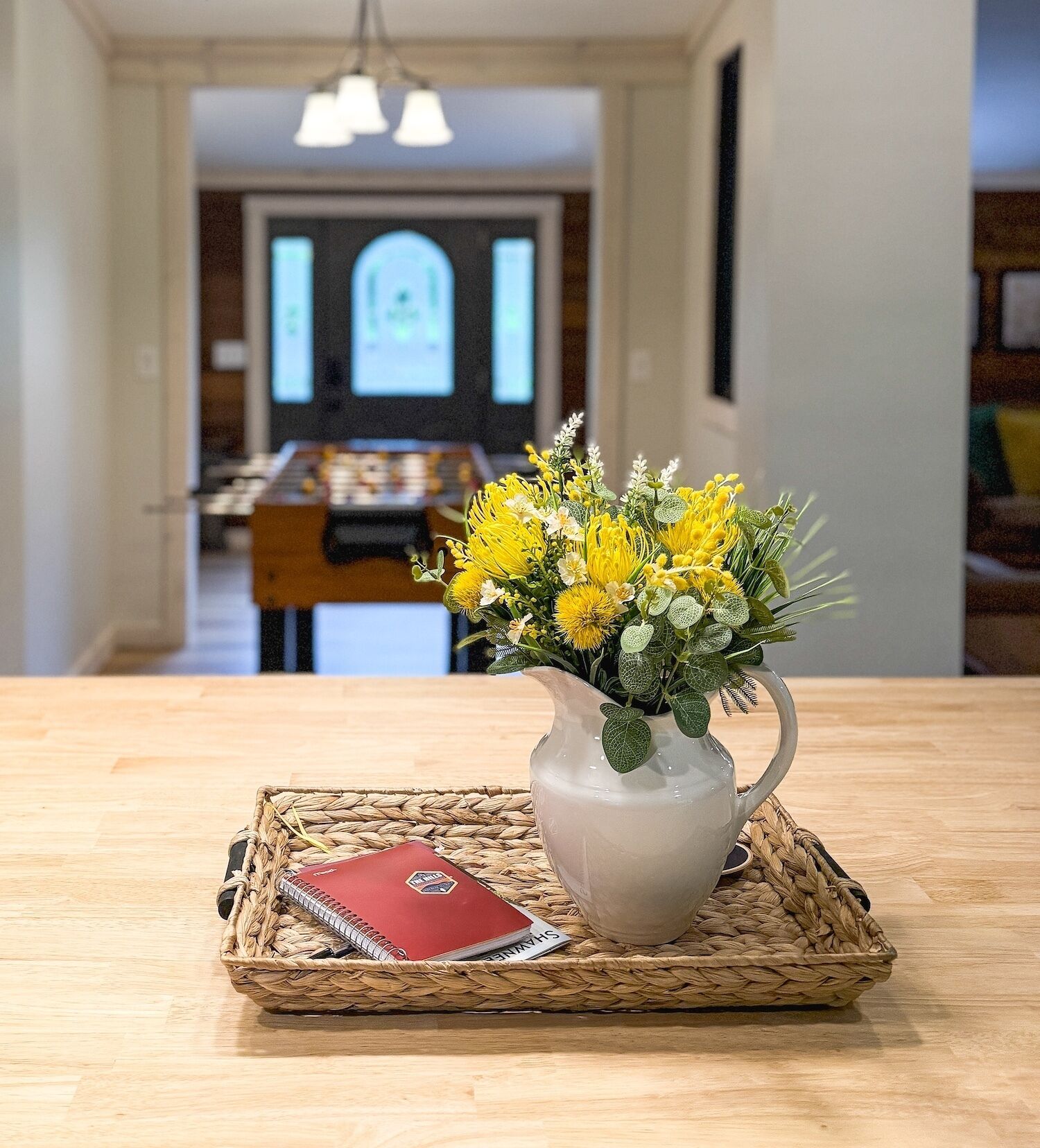 Une table en bois surmontée d’un set de table en fibres tressées, un pichet blanc orné de fleurs jaunes et un carnet rouge.