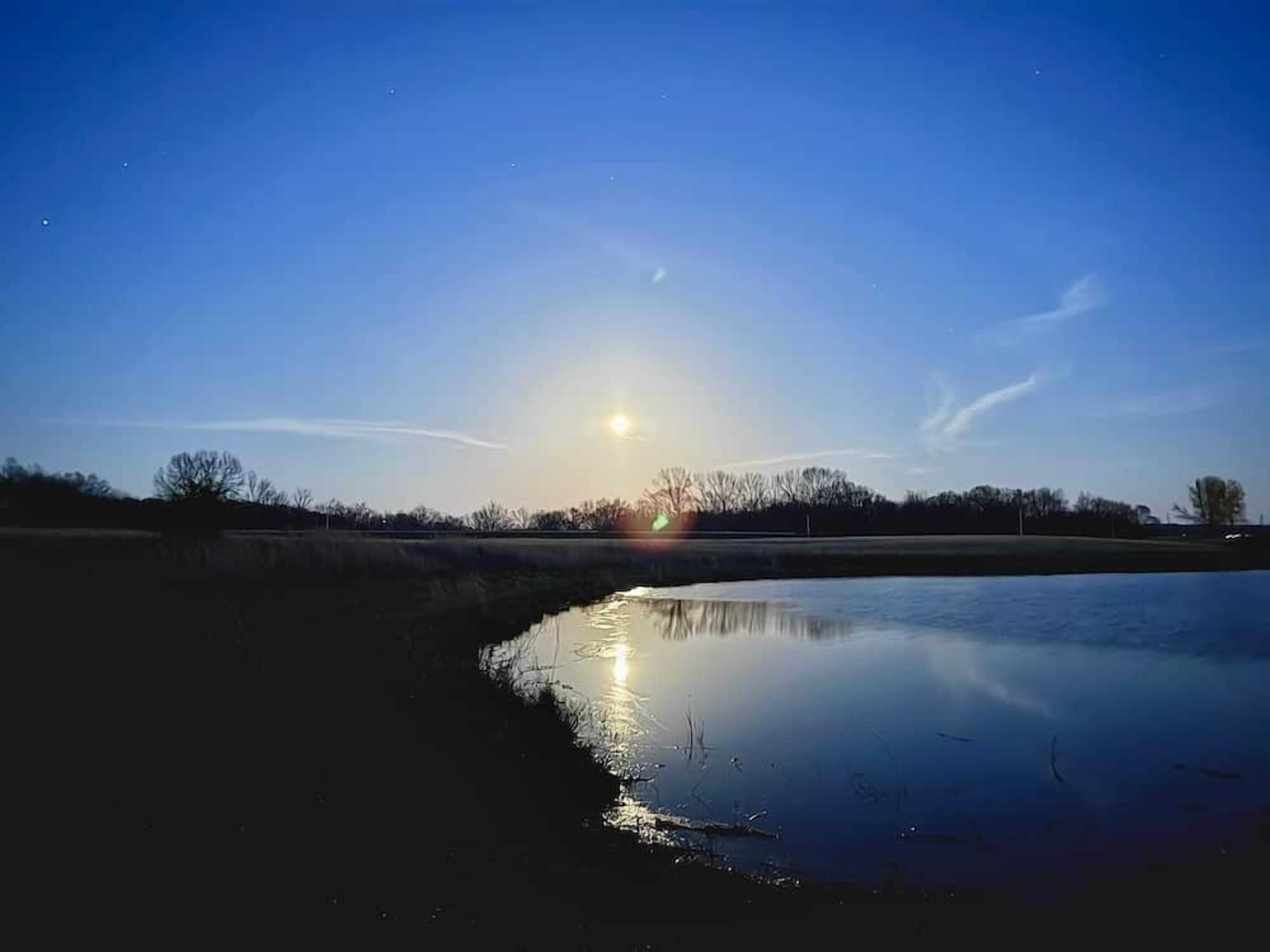 Ruhige Wasserfläche, die den Himmel und eine tief stehende, strahlende Sonne reflektiert.
