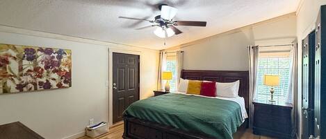 Une chambre à coucher avec un lit, un ventilateur de plafond, une porte, une table de chevet, une lampe et un tableau.