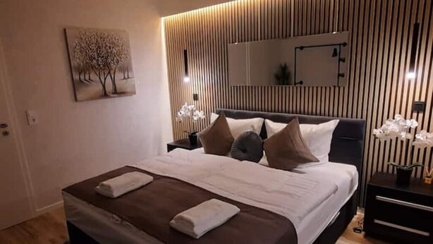 Een moderne slaapkamer met een groot bed, een nachtkastje, een spiegel en een wanddecoratie van bomen.