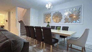 Une salle à manger avec une longue table, six chaises et un lustre. On aperçoit un escalier au fond de la pièce, qui mène à une autre pièce.