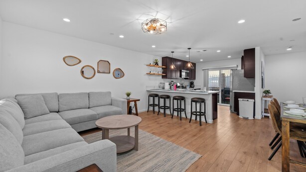 A modern living room with a grey sectional sofa, a round coffee table, and a kitchen area with a central island, bar stools, and wooden cabinets. - One World House with King Bed (Colton)