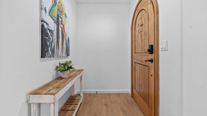 A hallway with a wooden door, a white bench, a potted plant, and a colorful abstract painting on the wall. - One World House with King Bed (Colton)
