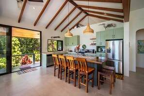 A modern kitchen with wooden beams, a large island with bar stools, and a view of lush greenery through the sliding glass door.