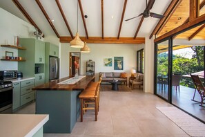 A modern kitchen with wooden beams, a dining area with a sofa, and a view of greenery through large sliding doors.