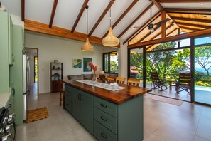 A spacious kitchen with a large island, wooden ceiling beams, and a view of lush greenery outside.