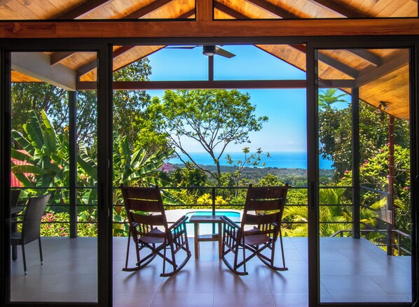 A wooden deck with two rocking chairs and a table, overlooking a lush landscape and the ocean.