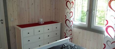 A bedroom with a wooden wall, a white dresser with drawers, a bed with a grey blanket, and a window with a view of trees.