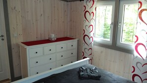 Une chambre avec un mur en bois, une commode blanche avec des tiroirs, un lit recouvert d’une couverture grise et une fenêtre donnant sur des arbres.