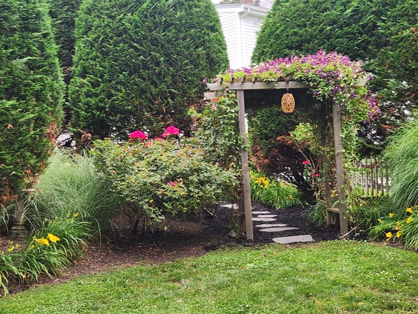 Un jardín con un porche de madera cubierto de plantas en flor, un camino de piedra y diversos arbustos y flores.