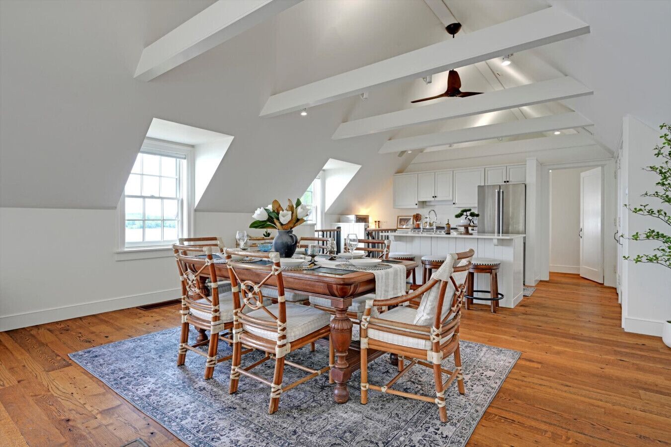 A spacious dining area with a wooden table and chairs, a kitchen in the background, and a ceiling fan.