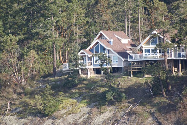 A two-story house with a large deck and a covered porch, surrounded by dense forest. - Waterfront Cabin at Currents, Otterbay, North Pender Island (Pender Island)
