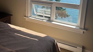 A bedroom with a large window overlooking a lake, a framed picture on the wall, and a bed with a gray blanket. - Waterfront Cabin at Currents, Otterbay, North Pender Island (Pender Island)