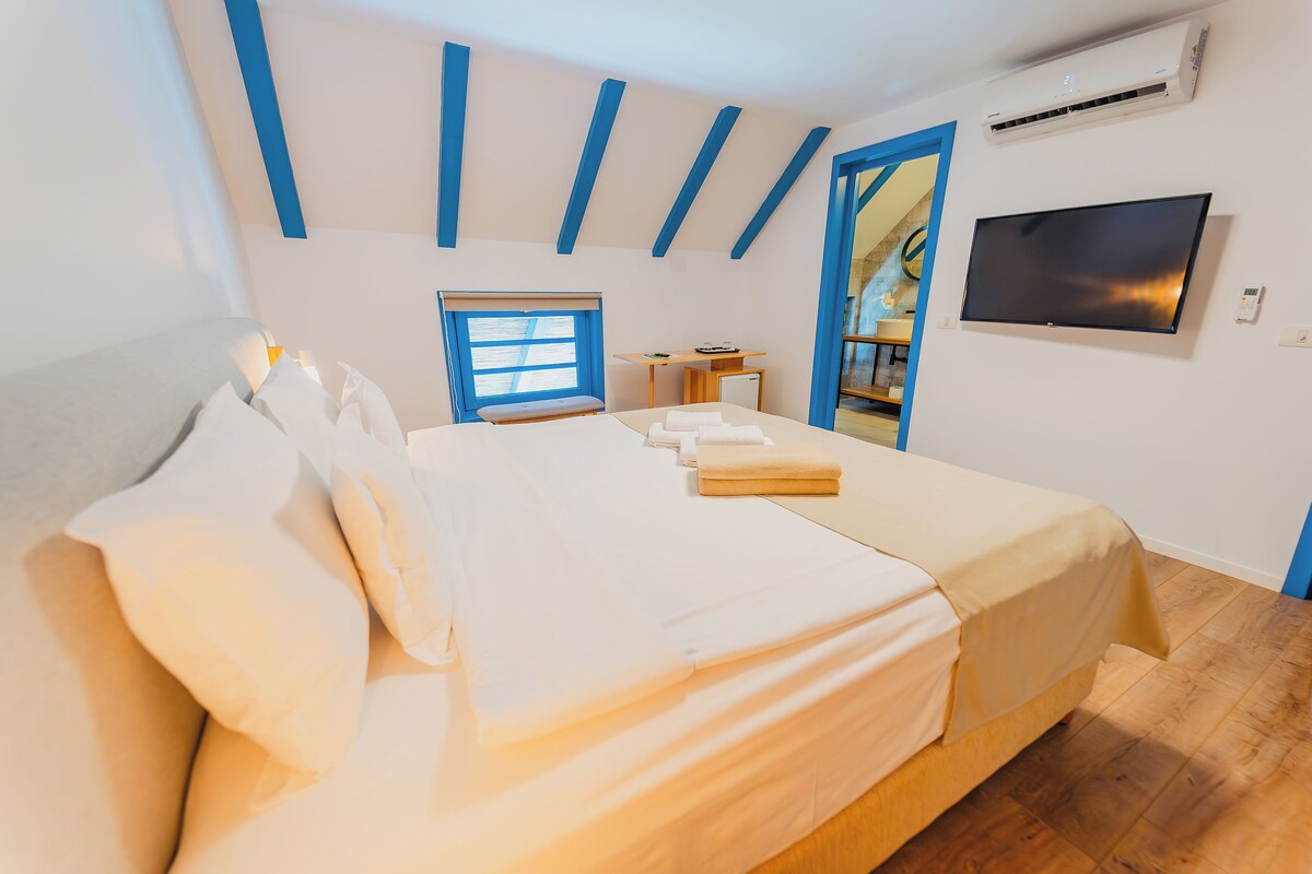 A bedroom with a bed, a desk, a TV, and a window.
