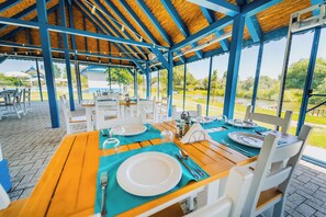 Un espace de restauration en plein air avec des tables prévues pour deux personnes, doté d’un toit de chaume et de grandes fenêtres offrant une vue sur le paysage environnant.