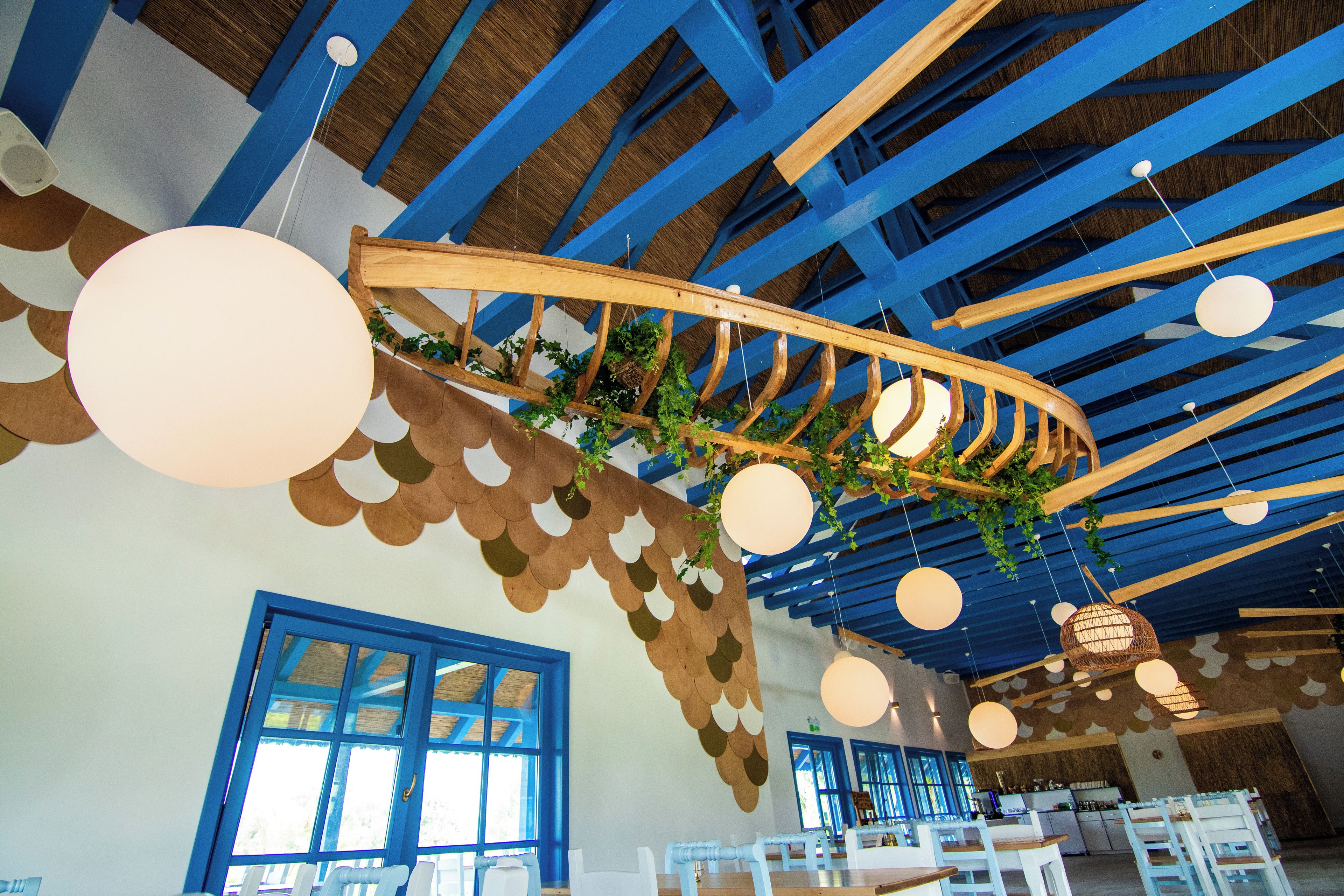 Ein Restaurantinnenraum mit blauen Deckenbalken, schwebenden, kugelförmigen Lampen und einer gewundenen Holzkonstruktion mit Pflanzen.