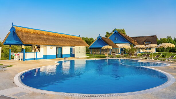 Une piscine avec un bâtiment à toit de chaume et un autre bâtiment avec un toit bleu.
