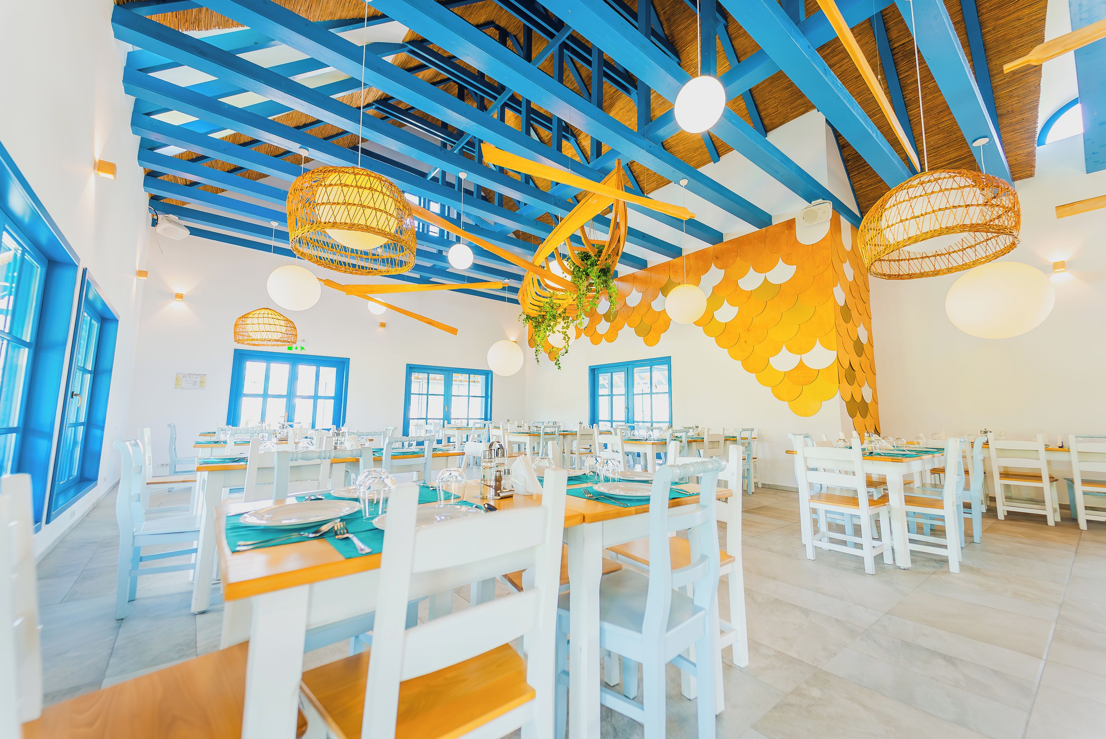 A dining area with white tables and chairs, blue beams on the ceiling, and hanging wicker lamps.