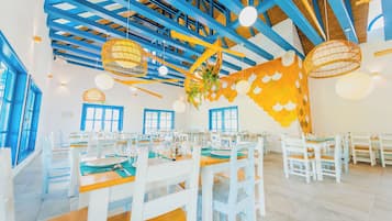 Un comedor con mesas y sillas blancas, vigas azules en el techo y lámparas de mimbre colgantes.