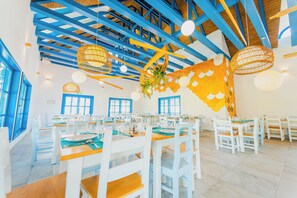 Zona pranzo con tavoli e sedie bianche, travi blu al soffitto e lampade di vimini appese.