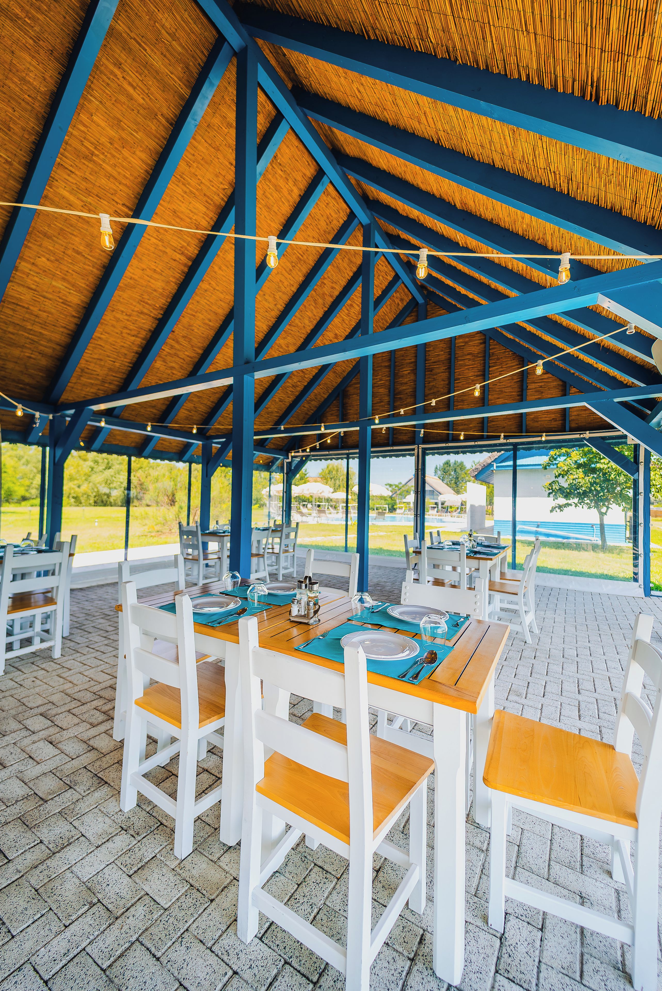An outdoor covered dining area with wooden tables and white chairs, featuring a view of the surrounding landscape.