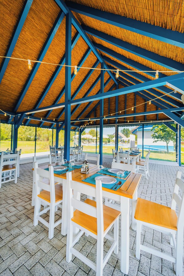 An outdoor covered dining area with wooden tables and white chairs, featuring a view of the surrounding landscape.