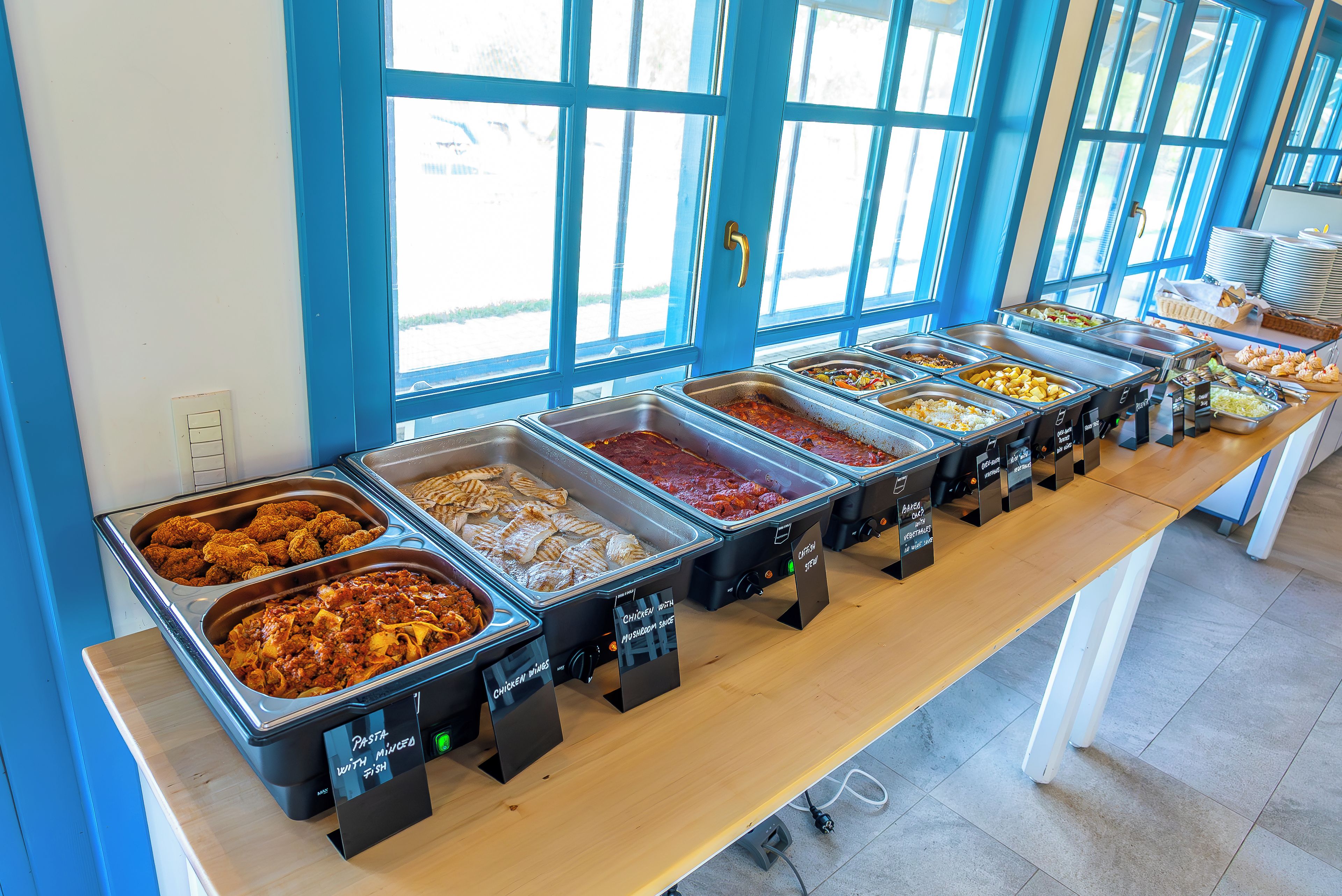 Un bufé con diversas opciones de comida, incluyendo pollo frito y pasta, dispuesto sobre una larga mesa con una pared azul y ventanas al fondo.