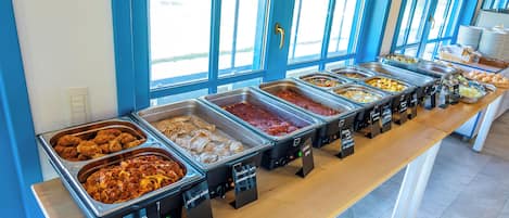 Un bufé con diversas opciones de comida, incluyendo pollo frito y pasta, dispuesto sobre una larga mesa con una pared azul y ventanas al fondo.