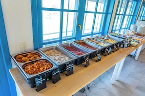 A buffet with various food options, including fried chicken and pasta, arranged on a long table with a blue wall and windows in the background. - Cherhana Resort (Crisan)