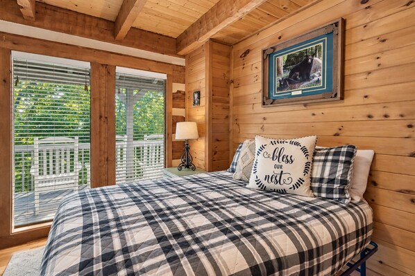 A wooden cabin bedroom with a bed, a framed picture on the wall, and a view through the window.