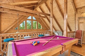 A wooden interior with a pool table, a hanging light fixture, and a window with a view of trees.