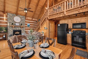 A wooden cabin interior with a dining area, kitchen, fireplace, and a staircase.